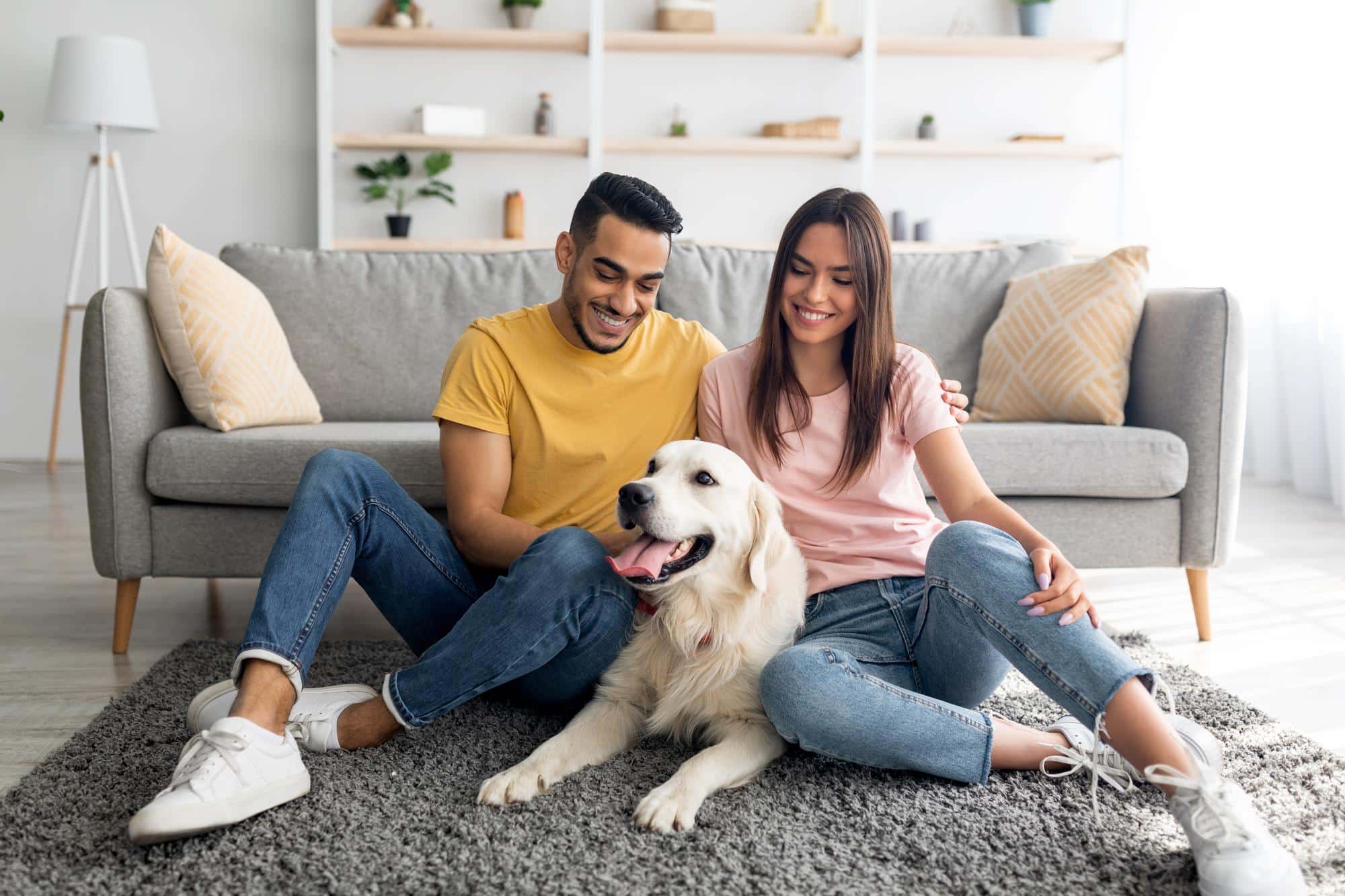 Full length portrait of cheery multiracial couple with their pet dog sitting on floor at home Friends drinking